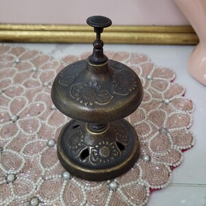 Puede incluir: Una campana antigua de color bronce con detalles florales. La campana tiene una parte superior en forma de c&uacute;pula y una base circular con recortes decorativos. La campana est&aacute; colocada sobre un mantel individual rosa con cuentas y borde festoneado. Es un objeto decorativo.