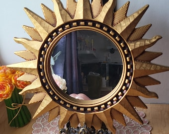 May include: Gold-colored sunburst mirror with an oval mirror center. The frame has textured rays and a dark brown border. The mirror is supported by a decorative metal stand and sits on a pink lace doily.