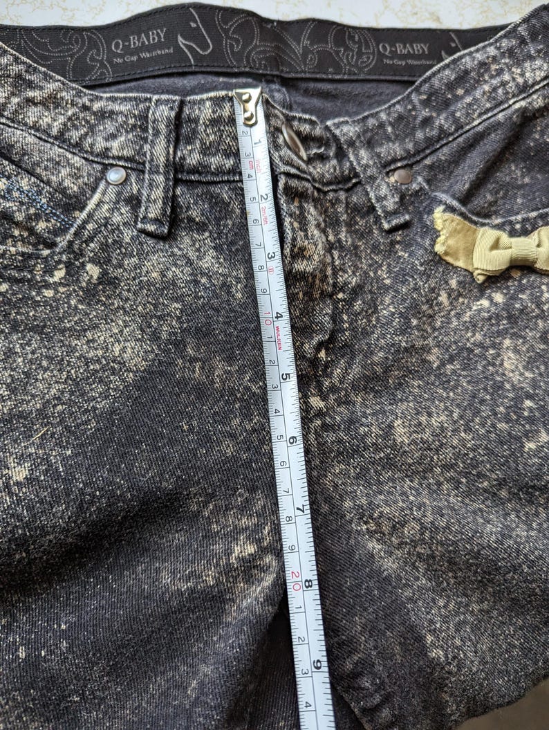May include: Black acid wash denim jeans with a yellow bow detail. The jeans have a measurement tape running vertically along the side, measuring 9 inches.