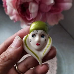 K&ouml;nnte beinhalten: Eine wei&szlig;e Keramikfigur eines Frauenkopfes mit gr&uuml;nen und schwarzen Details. Die Figur tr&auml;gt einen gr&uuml;nen Hut und hat blaue Augen und rote Lippen.