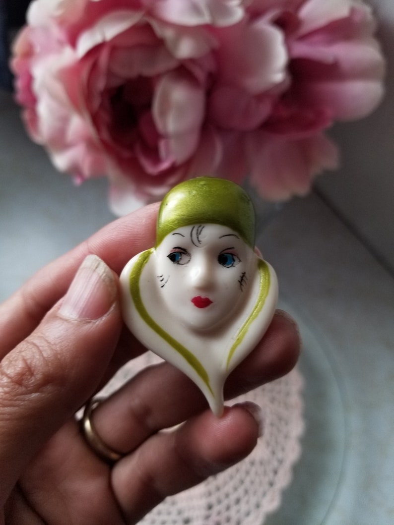 K&ouml;nnte beinhalten: Eine wei&szlig;e Keramikfigur eines Frauenkopfes mit gr&uuml;nen und schwarzen Details. Die Figur tr&auml;gt einen gr&uuml;nen Hut und hat blaue Augen und rote Lippen.