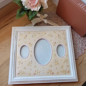 Può includere: Una cornice bianca con tre aperture ovali e un motivo floreale. La cornice ha uno sfondo color crema con fiori rosa e gialli in rilievo. Un bouquet di fiori rosa e bianchi è nelle vicinanze.