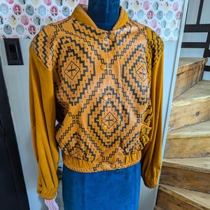 Op de afbeelding: Een mosterdgele leren jas met een zwart geometrisch patroon. De jas heeft een ritssluiting en een geribbelde gebreide kraag, manchetten en tailleband.