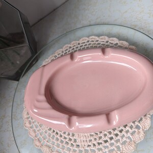 May include: A pink ceramic ashtray with a decorative design. The ashtray is sitting on a white doily.