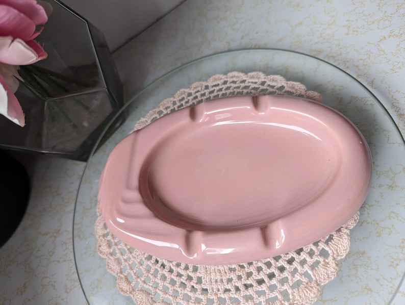 May include: A pink ceramic ashtray with a decorative design. The ashtray is sitting on a white doily.