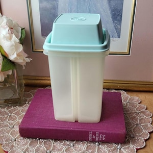 May include: A vintage, rectangular food storage container with a light blue lid. The container is translucent with a vertical divider. It sits on a purple book. The background includes a framed picture and a vase of white flowers.