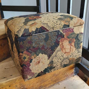 May include: A floral patterned storage ottoman with a hinged lid. The ottoman is made of a fabric with a variety of colors, including beige, brown, red, and green.