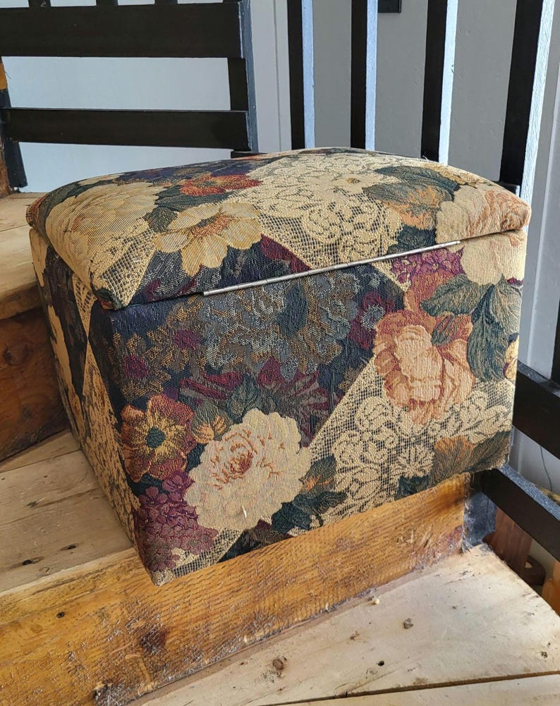 May include: A floral patterned storage ottoman with a hinged lid. The ottoman is made of a fabric with a variety of colors, including beige, brown, red, and green.