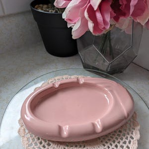 May include: A pale pink ceramic ashtray with a rounded oval shape. The ashtray is sitting on a white crocheted doily.