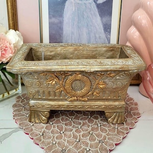 Puede incluir: Una jardinera rectangular dorada con detalles ornamentados, incluido un diseño central de corona. La jardinera se asienta sobre cuatro pies decorativos y se coloca sobre un tapete con cuentas estampadas. Un retrato enmarcado es visible en el fondo.