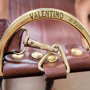 May include: Close-up of a brown leather bag with a gold metal clasp. The clasp is engraved with the words "VALENTINO di Paolo".