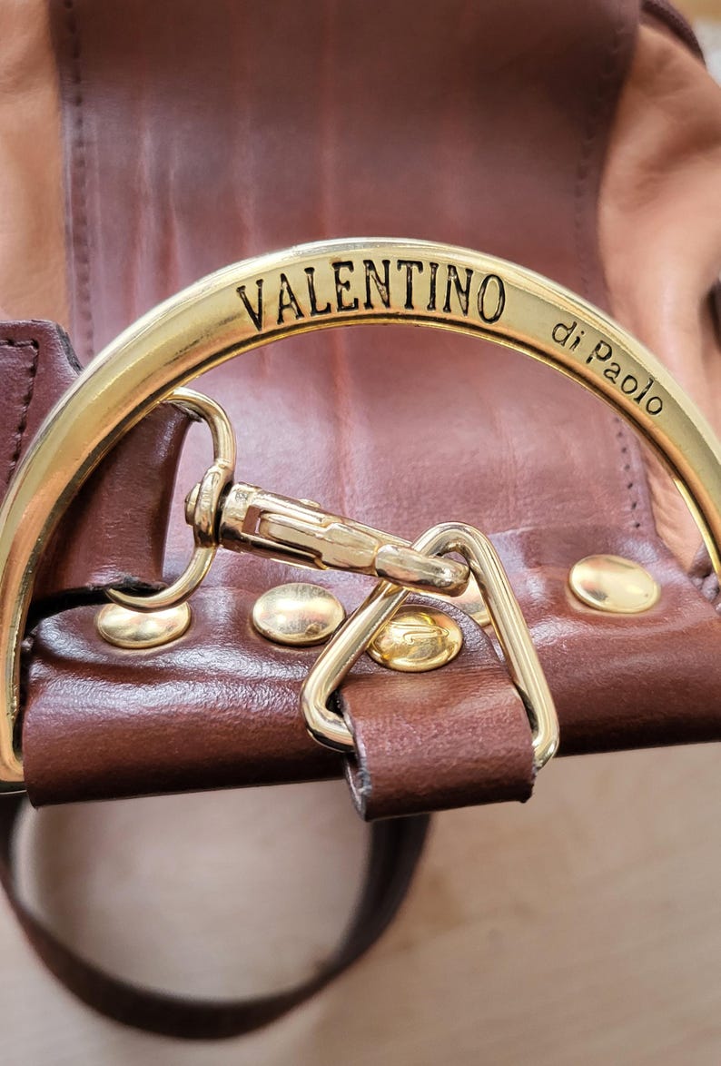 May include: Close-up of a brown leather bag with a gold metal clasp. The clasp is engraved with the words "VALENTINO di Paolo".