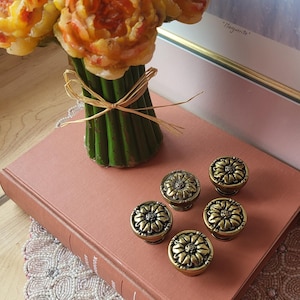 Peut inclure: Un bouquet de fleurs artificielles jaunes et oranges dans un vase vert noué avec de la ficelle est posé sur un livre rose poussiéreux. Cinq boutons anciens en laiton en forme de tournesol sont disposés sur le livre, avec un napperon en dentelle perlée.