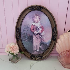 May include: Oval framed portrait of a young child holding a small dog. The portrait has a gold-toned frame with ornate detailing. The child is wearing a red jacket and pink pants. A small bouquet of pink flowers sits in a glass vase.