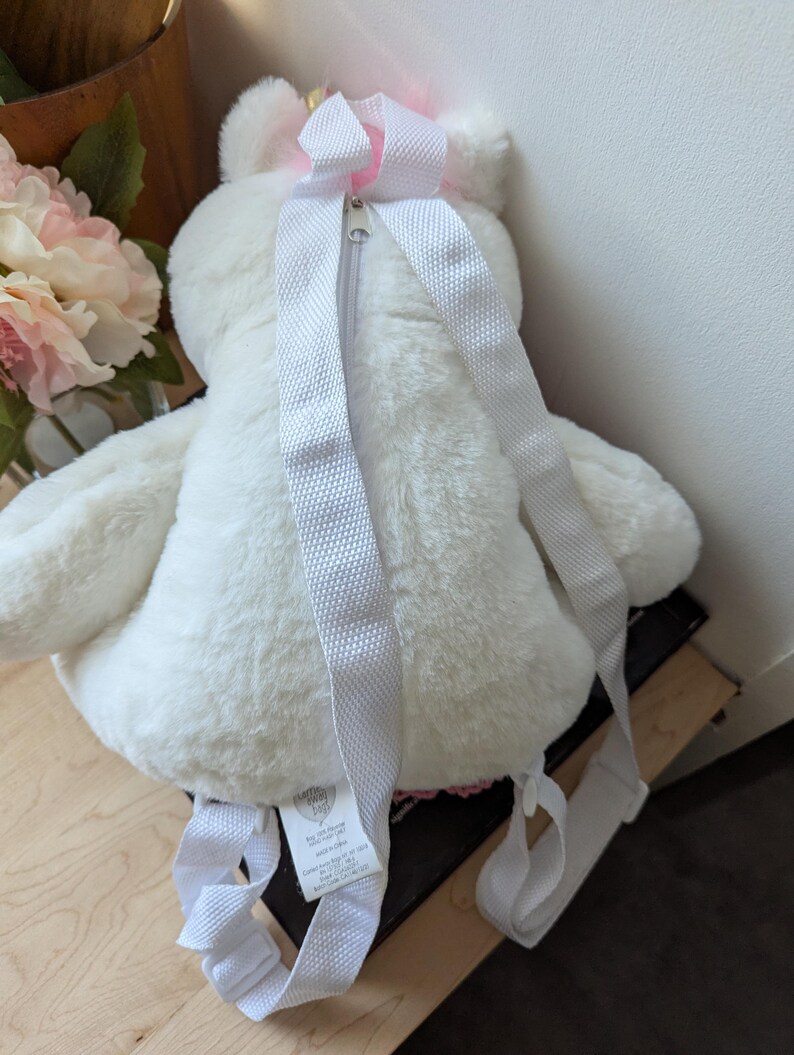 May include: White plush unicorn backpack with pink accents and a zipper closure. The backpack has adjustable straps.