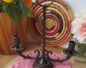 Puede incluir: Un candelero de metal negro con tres brazos y un asa de anillo. El candelero está sentado sobre un encaje de ganchillo rosa.