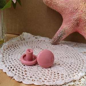 Pode incluir: Dois botões de gaveta de plástico rosa com formato redondo e um pequeno caule. Os botões estão sobre um crochê branco.