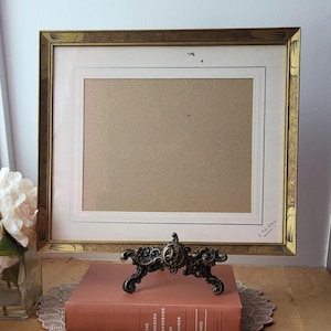 Puede incluir: Un soporte decorativo de metal color bronce con un diseño ornamentado, apoyado sobre un libro color coral. Una imagen enmarcada con borde dorado está en el fondo. El libro está sobre un tapete de encaje.