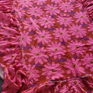 Puede incluir: Una tela rosa con estampado floral con grandes flores tipo margarita en tonos de rosa, morado y amarillo. La tela tiene un borde con volantes. El fondo es de color marrón rojizo.