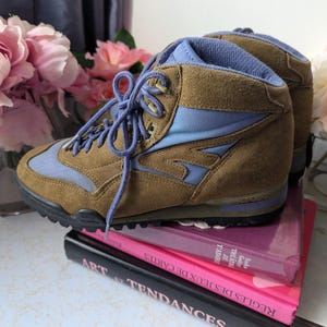 May include: Brown suede hiking boots with blue accents and laces. The boots are sitting on top of a stack of books.