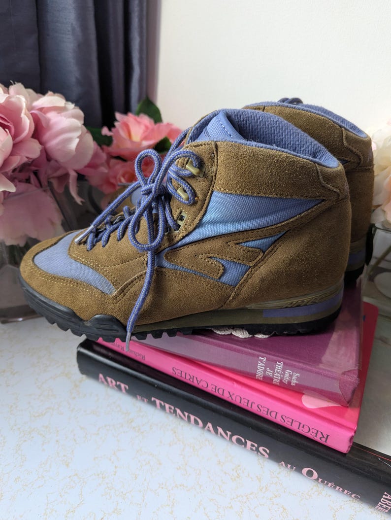 May include: Brown suede hiking boots with blue accents and laces. The boots are sitting on top of a stack of books.