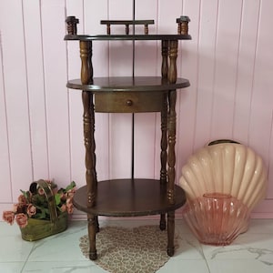 Puede incluir: Una mesa auxiliar de madera marrón oscuro de tres niveles con un pequeño cajón. La mesa tiene patas torneadas y una parte superior decorativa. Un jarrón rosa en forma de concha y un jarrón de cristal rosa transparente están junto a la mesa.