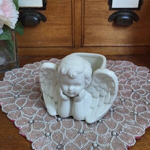 Peut inclure: Un pot de fleurs en céramique crème en forme d'ange, avec des ailes détaillées et une expression sereine. Il repose sur un dessous de plat fleuri et perlé. En arrière-plan, un meuble en bois.