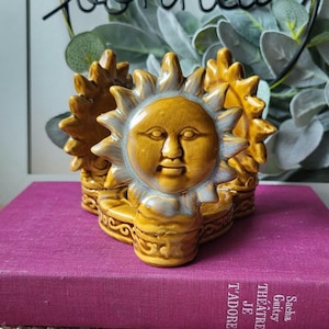 Puede incluir: Un servilletero de cerámica color amarillo mostaza con un diseño tridimensional de cara de sol con rayos radiantes. La base presenta un patrón decorativo de volutas en relieve, y el motivo del sol se repite en ambos lados de forma simétrica.