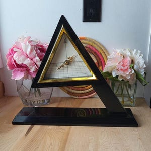 May include: A black triangular clock with gold accents and a white face. The clock is on a black stand and is surrounded by flowers.