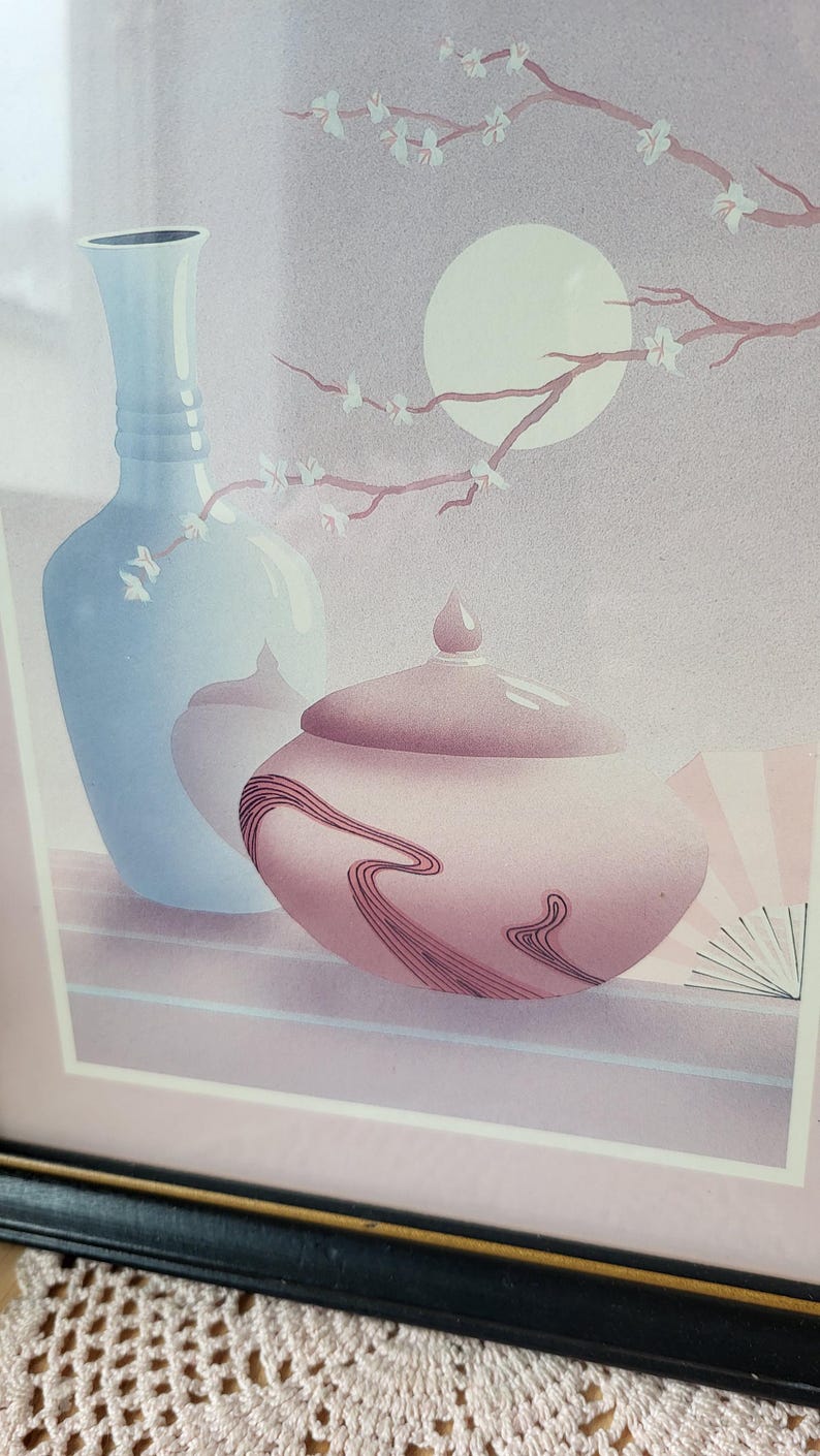 Op de afbeelding: Een ingelijste print van een stilleven met een blauwe vaas, een roze dekselpot met een draaiend ontwerp en een tak met witte bloemen tegen een lichtpaarse achtergrond.