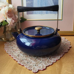 May include: A navy blue enamel teapot with a gold-colored knob and trim. The teapot has a black handle and a spout. It sits on a decorative doily with a red border. A framed picture and flowers are in the background.