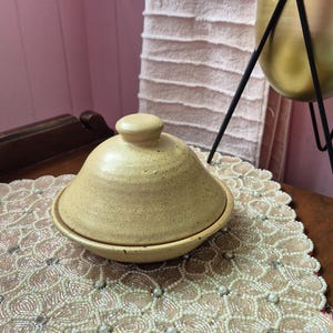 May include: A round, speckled, light yellow ceramic lidded dish with a conical top and a small knob handle. The dish features a smooth, matte-like finish with subtle dark flecks throughout the clay body, resting on a flat, circular base.