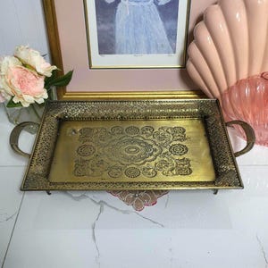 May include: A rectangular, antique brass tray with ornate floral and scrollwork designs. The tray has two curved handles and a decorative border. The tray is displayed on a white surface, with a framed picture and pink seashell in the background.