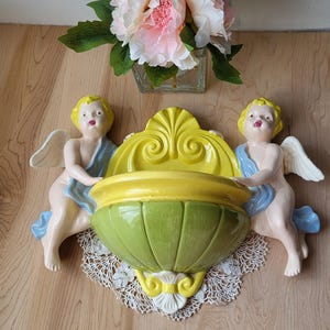May include: A vintage wall-mounted planter in green, yellow, and white. Two cherubic figures with yellow hair and white wings flank the planter. The planter is decorated with a lace doily and a bouquet of pink and white flowers.