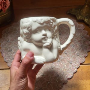 May include: White ceramic mug featuring a cherub design. The mug has a detailed relief of a cherub's face and body, with a curved handle. The mug is held in a hand, and the background includes a beaded placemat and wooden surface.