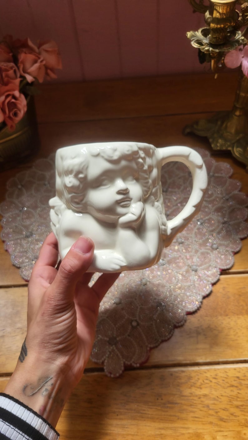 May include: White ceramic mug featuring a cherub design. The mug has a detailed relief of a cherub's face and body, with a curved handle. The mug is held in a hand, and the background includes a beaded placemat and wooden surface.