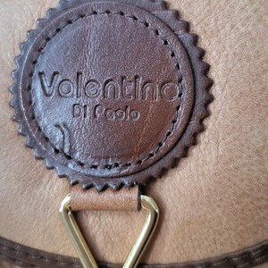 May include: Close-up of a brown leather purse with a gold-colored metal clasp. The purse has a brown leather tag with the text "Valentino Di Paolo" embossed in gold.