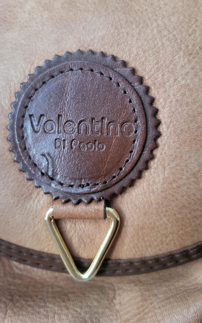 May include: Close-up of a brown leather purse with a gold-colored metal clasp. The purse has a brown leather tag with the text "Valentino Di Paolo" embossed in gold.