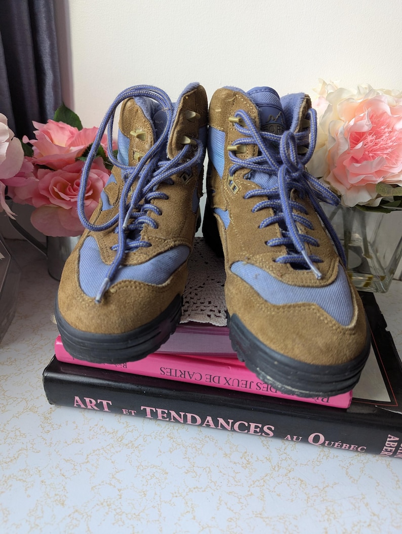 May include: A pair of brown and blue suede hiking boots with laces. The boots feature a light blue nylon panel on the side and a brown suede upper. The boots are sitting on top of a stack of books.