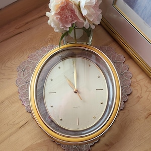 May include: A gold-framed quartz clock with a cream face and gold hands. The clock is on a decorative doily, with a vase of pink flowers and a framed picture in the background. The clock face reads "QUARTZ".