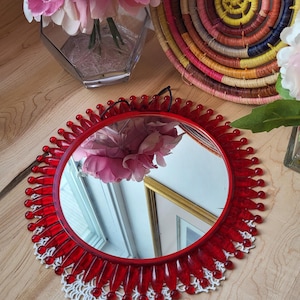 以下が含まれることがあります： ビーズの縁取りと白いレースのトリムが付いた赤い化粧鏡。鏡にはピンクの牡丹の花束が映っています。