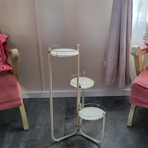 Op de afbeelding: Een plantenstandaard met drie niveaus, met een crèmekleurig frame en witte ronde planken. Elke plank heeft een goudkleurige metalen rand. De standaard staat voor een roze muur en een raam met gordijn.