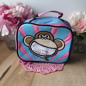 May include: A blue and pink lunch bag with a monkey face design. The monkey has a zipper for a mouth and a banana in its hand. The text "Original Banana" is printed on the bag.
