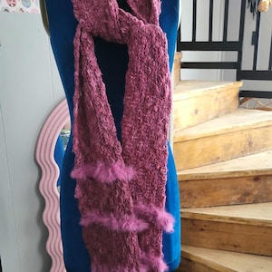 May include: A pink knitted scarf with a fluffy trim. The scarf is displayed on a mannequin.