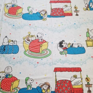 Puede incluir: Una tela blanca con un patrón repetido de Snoopy y sus amigos durmiendo en varias camas. La tela presenta una combinación de colores azul, rojo y amarillo con estrellas blancas sobre un fondo azul claro.