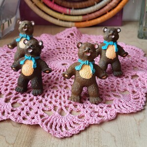 May include: Four brown teddy bears with blue ribbons around their necks stand on a pink crocheted doily. The bears are all facing forward and have their mouths open in a smile.