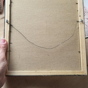 Op de afbeelding: De achterkant van een houten fotolijst met een zwarte rand. De lijst heeft een draadbeugel die aan de achterkant is bevestigd.