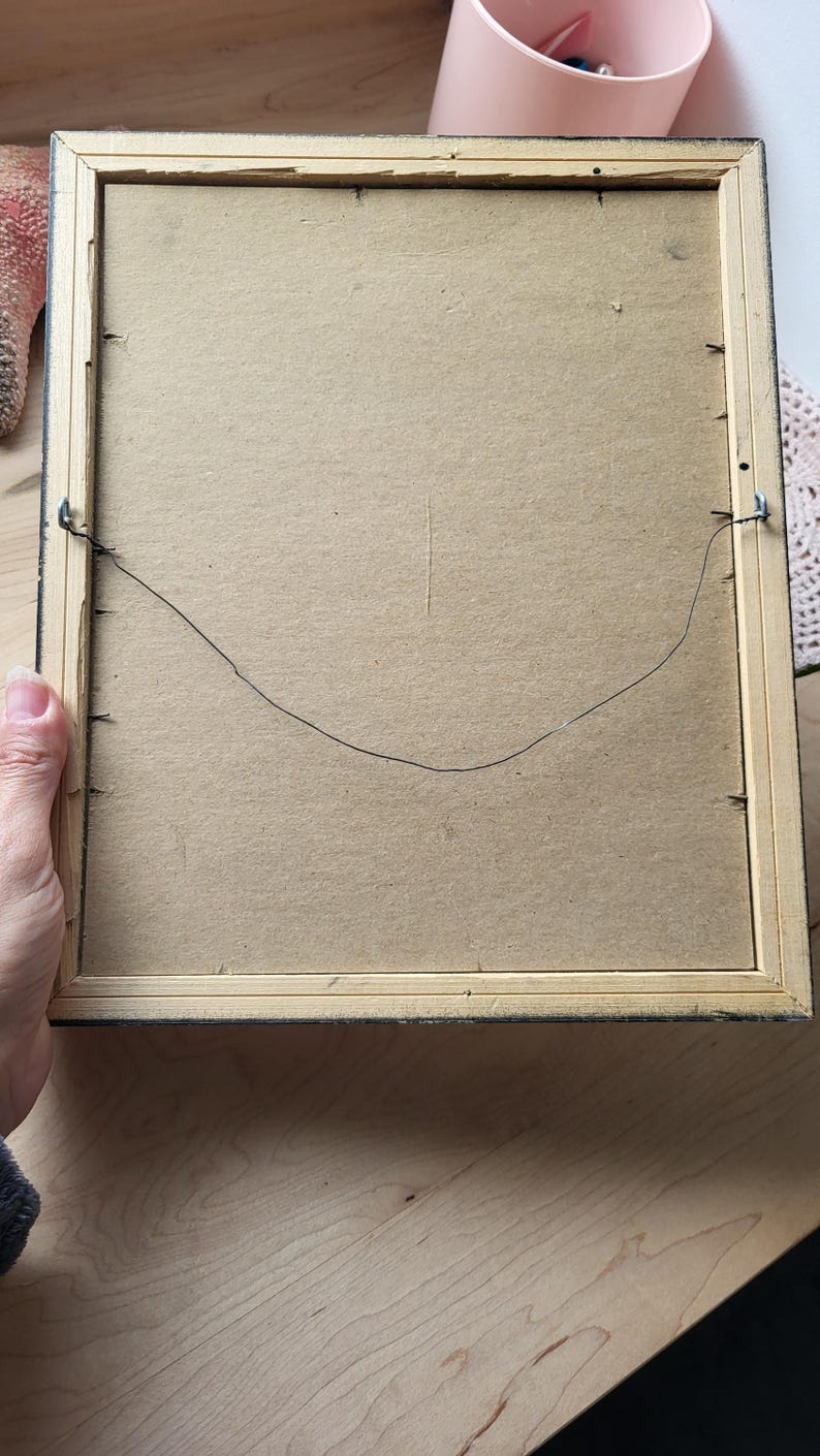 Op de afbeelding: De achterkant van een houten fotolijst met een zwarte rand. De lijst heeft een draadbeugel die aan de achterkant is bevestigd.