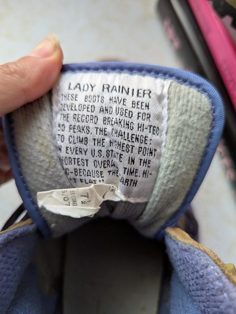May include: A close-up of a blue and white label with the text "Lady Rainier" and a description of the boots' features. The text reads: "These boots have been developed and used for the record breaking hi-tec 50 peaks. The challenge: to climb the highest point in every U.S. state in the shortest overall time. Hi-tech flat earth."