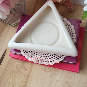 May include: White ceramic triangle shaped dish with a raised rim. The dish is sitting on top of two books, one purple and one red. A white crocheted doily is underneath the dish.